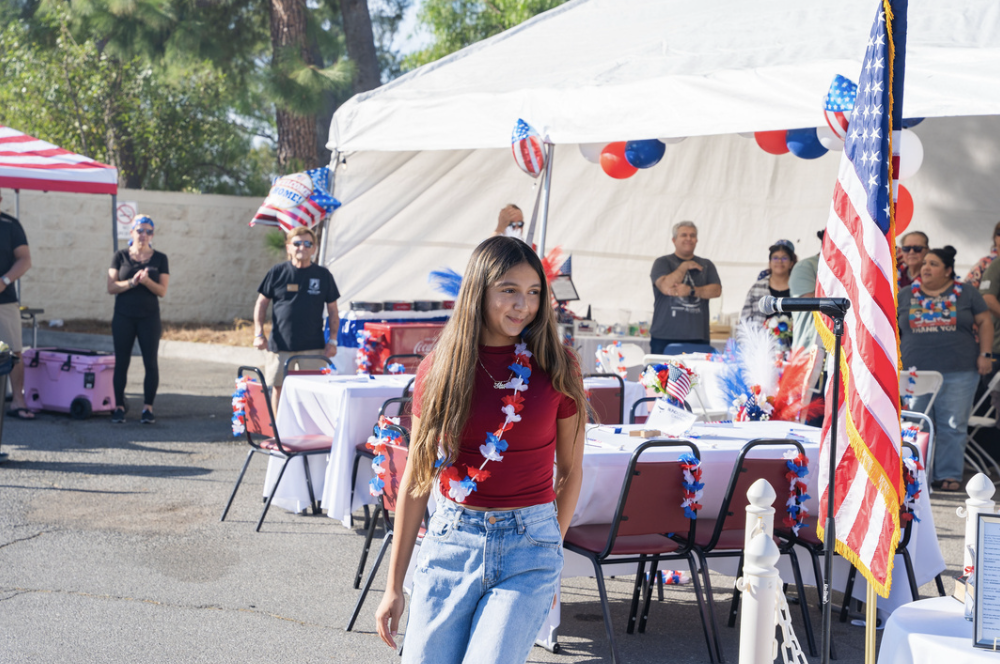 A young woman wearing a red shirt and jeans stands in front of a decorated outdoor event with American flags, patriotic streamers, and balloons. Several people are gathered around tables and tents, also decorated with patriotic themes.