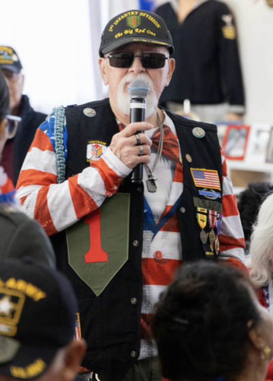 An elderly man wearing sunglasses, a black cap with yellow lettering, and a vest decorated with patches and medals, holding a microphone. Several other veterans are visible in the background, indicating a veterans' event.