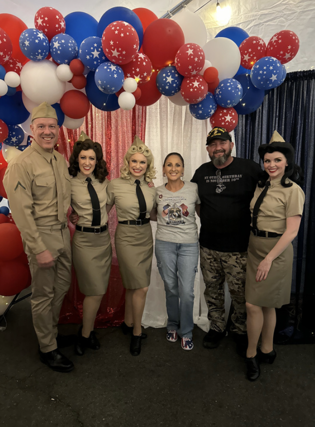 Group of six people dressed in vintage military costumes and casual clothing standing under colorful patriotic balloon arch, celebrating a patriotic or military-themed event.