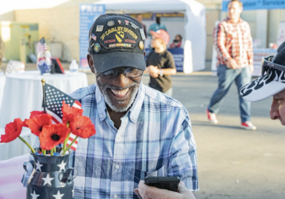 Smiling man wearing a cap with military patches at an outdoor event, looking at his phone, with a vase of red flowers and American flags in front of him.
