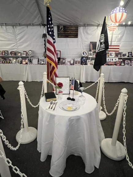 A dining table set for one with a white tablecloth, a plate with a fried egg, utensils, a glass of water, a small American flag, and a rose. There are American and military flags behind, and a display of framed photos and memorabilia in the backgroun