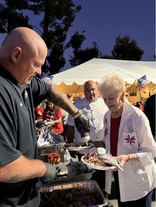 A man serving food at an outdoor event with a woman holding a plate, under a white canopy with American flags, with several people in the background.