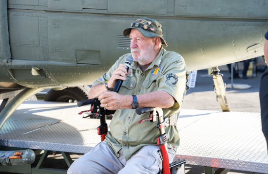 An elderly man in military attire sits in a wheelchair, holding a microphone, in front of a military helicopter during an event.