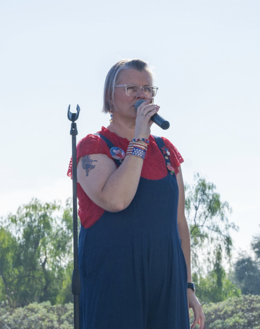 A woman with short gray hair and glasses holding a microphone, wearing a red top with lace details and a dark blue dress with patriotic star and stripe accessories, standing outdoors with trees and a cloudy sky in the background.