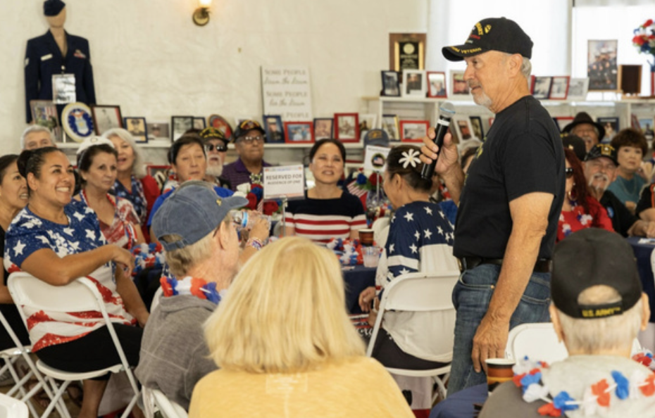 A man in a black t-shirt and cap addresses a crowd of veterans and seniors at an event celebrating patriotism, with many wearing American flag clothing, in a decorated room with photos and memorabilia.