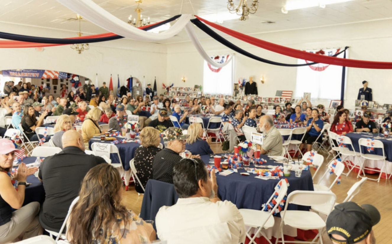 A patriotic celebration with people seated at decorated tables featuring red, white, and blue theme, patriotic banners and flags, and display cases with military uniforms in a large hall.