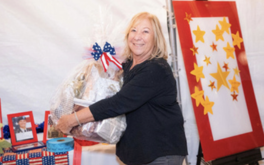 A woman with blonde hair smiling and hugging a large gift wrapped in silver paper with a red, white, and blue bow. There are framed photos and patriotic decorations in the background, including a red, white, and blue tablecloth and a red display boar