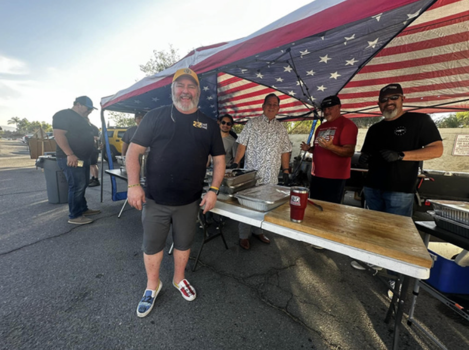 Group of five people standing under a canopy with an American flag pattern, smiling at the camera, at an outdoor event or gathering.