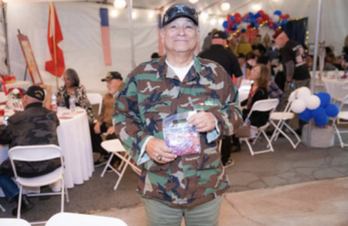 A smiling man in camouflage jacket and cap holding a gift, standing inside a decorated event tent with other people, tables, and balloons.