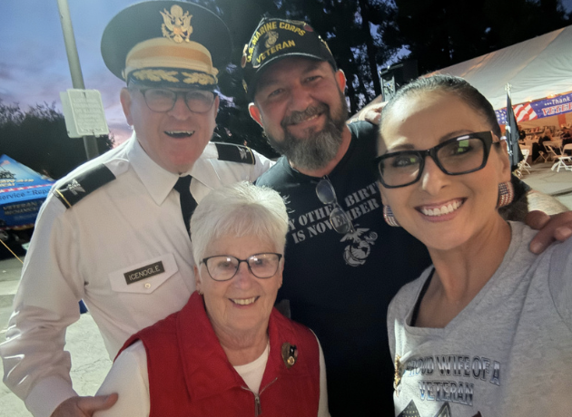 Four smiling people taking a selfie at an outdoor event during the evening. One man is dressed in a pilot's uniform with a captain's hat, another man is wearing a black cap and t-shirt, a woman is wearing glasses and a gray shirt, and an elderly woma