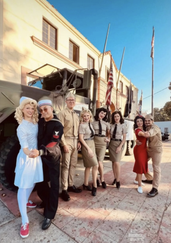 People dressed in vintage military and pin-up style costumes posing in front of a military vehicle with flags, in a themed event setting.