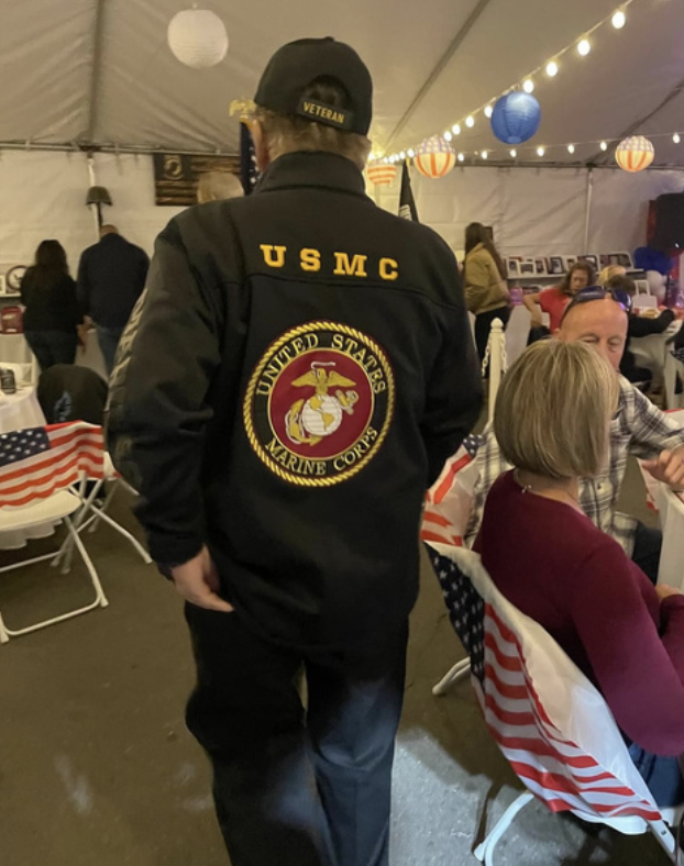 Man wearing US Marine Corps jacket with Marine emblem, and a cap labeled 'Veteran,' at a festive event with American flags and autumn-themed decorations.