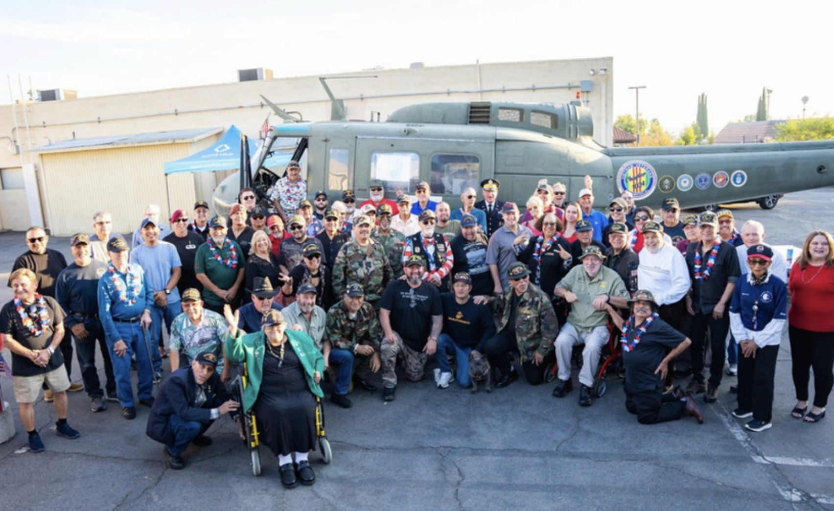 Group of veterans and civilians posing in front of a military helicopter during an outdoor event.