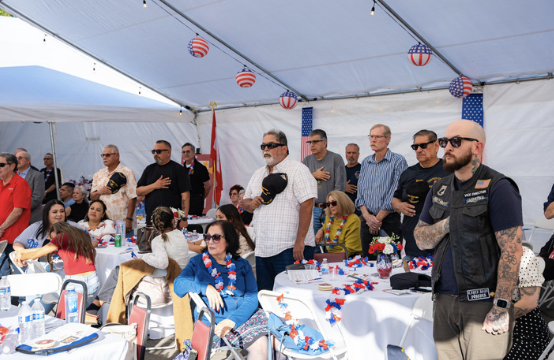 People standing with hats in hand during an outdoor Independence Day celebration under a decorated tent.
