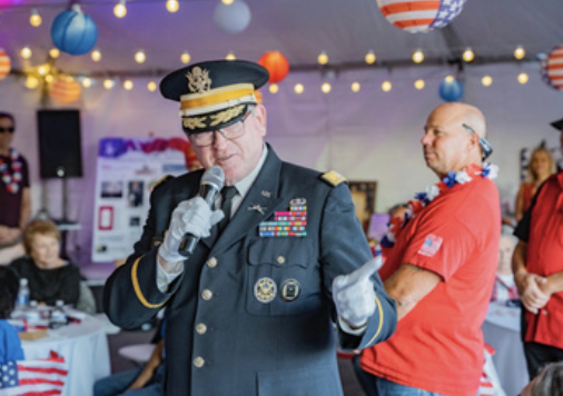 A man in a military uniform speaking into a microphone at a patriotic celebration, with people and American-themed decorations in the background.