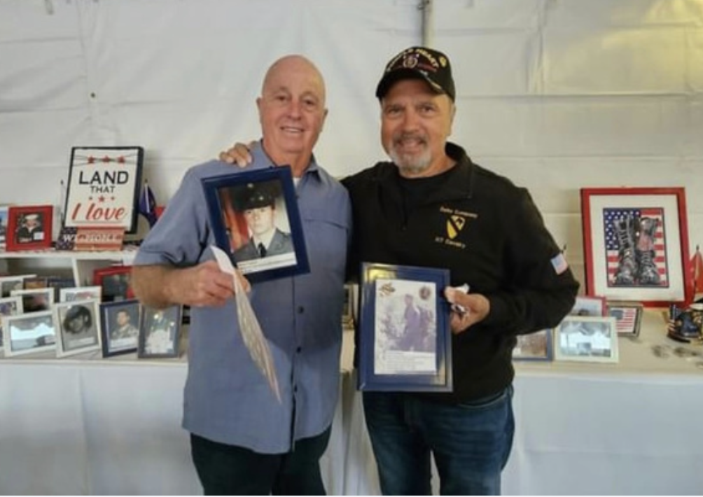 Two men standing indoors, smiling, holding framed photographs of military personnel. The background has patriotic decorations and a display of framed photos and memorabilia.