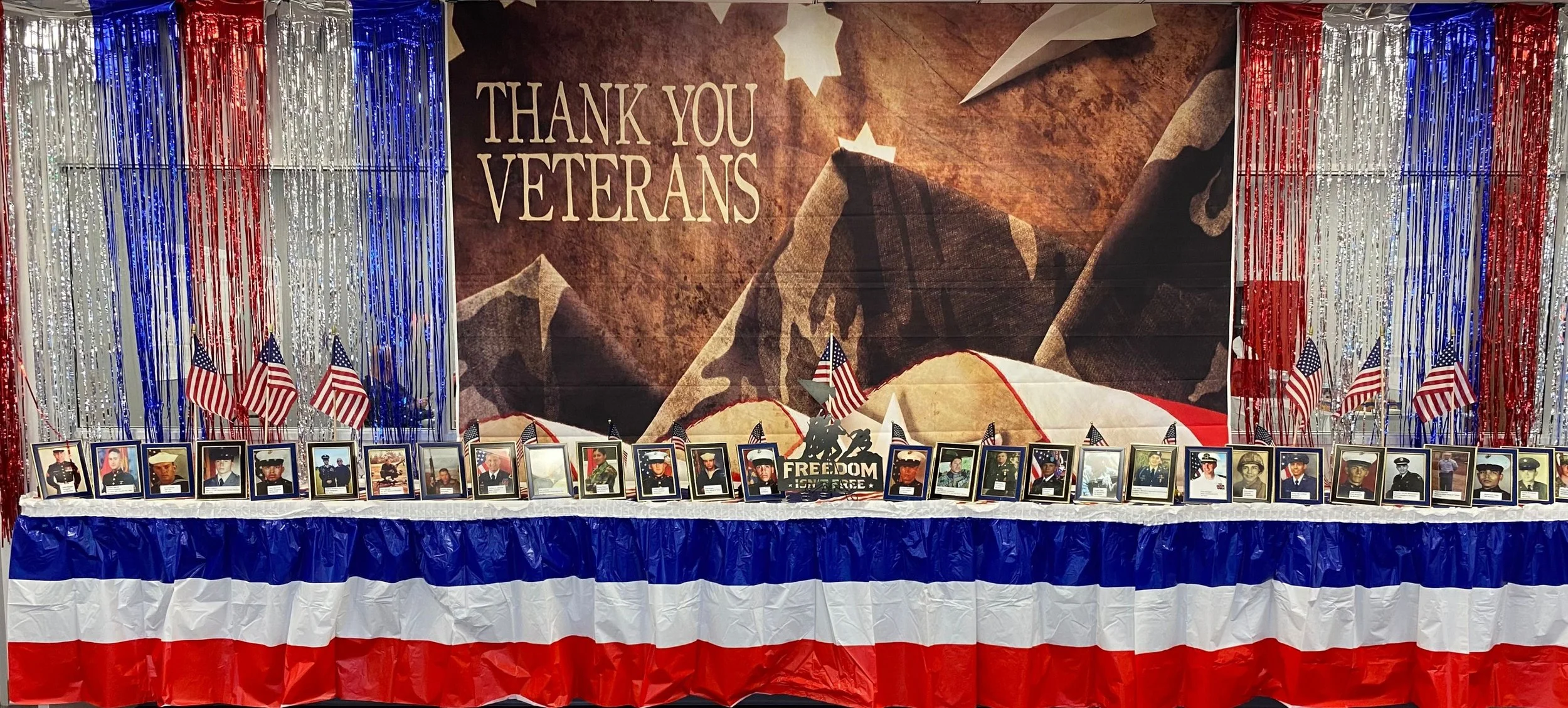 Display honoring veterans with framed photographs of servicemen and women arranged on a table, American flags, and red, white, and blue decorations, including a large banner that says 'Thank You Veterans'.