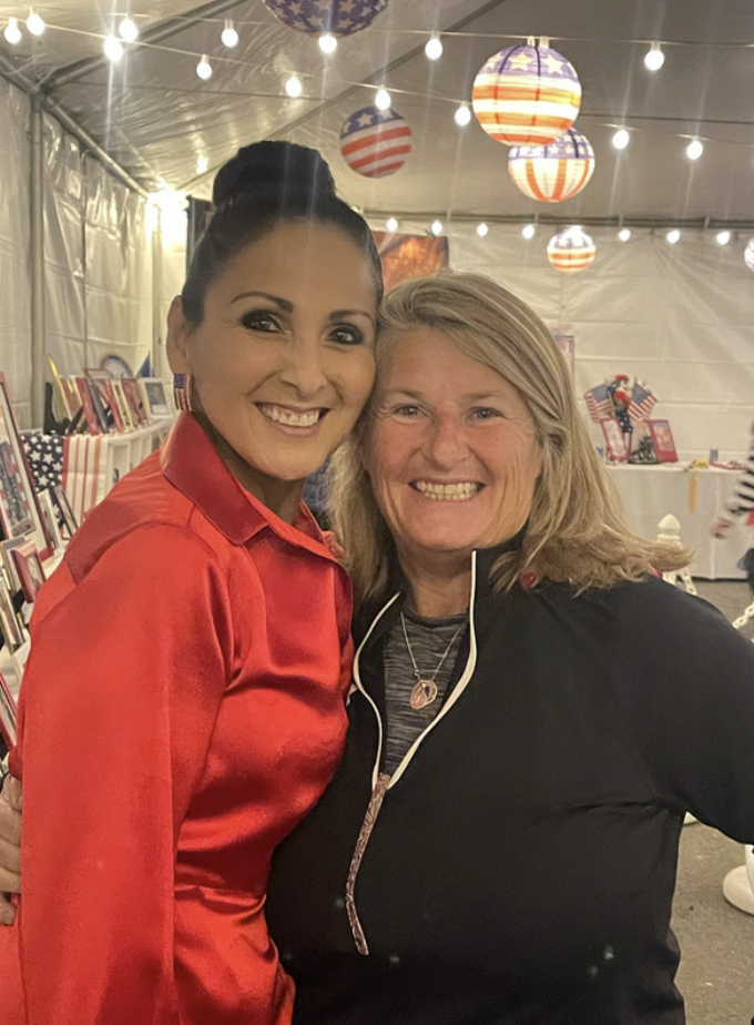Two women smiling and posing together at an event decorated with American flags and patriotic decorations, with string lights hanging overhead.