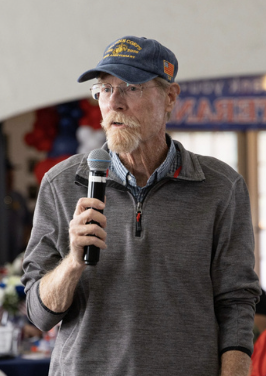 An older man with glasses and a beard, wearing a blue cap and gray quarter-zip pullover, holding a microphone and speaking at an indoor event.
