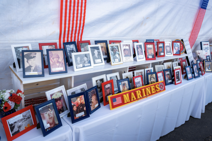 Display of framed photographs of military personnel, arranged on tables with a Marine Corps sign, decorated with American flags and flowers, likely at a memorial or tribute event.