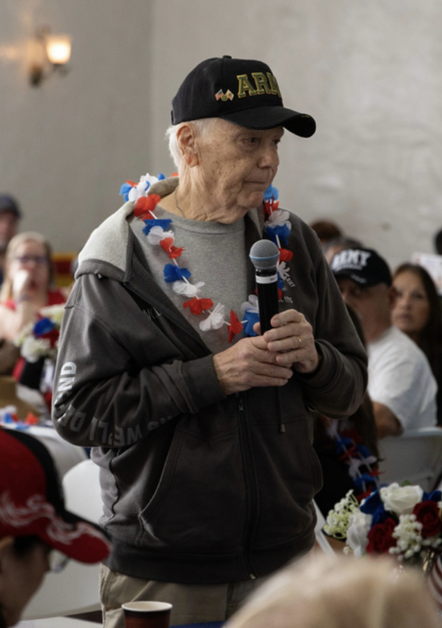 An elderly man wearing a black hat with gold lettering, a gray hoodie, and a patriotic flower lei, holding a microphone during a gathering with other people, some wearing patriotic hats and accessories.
