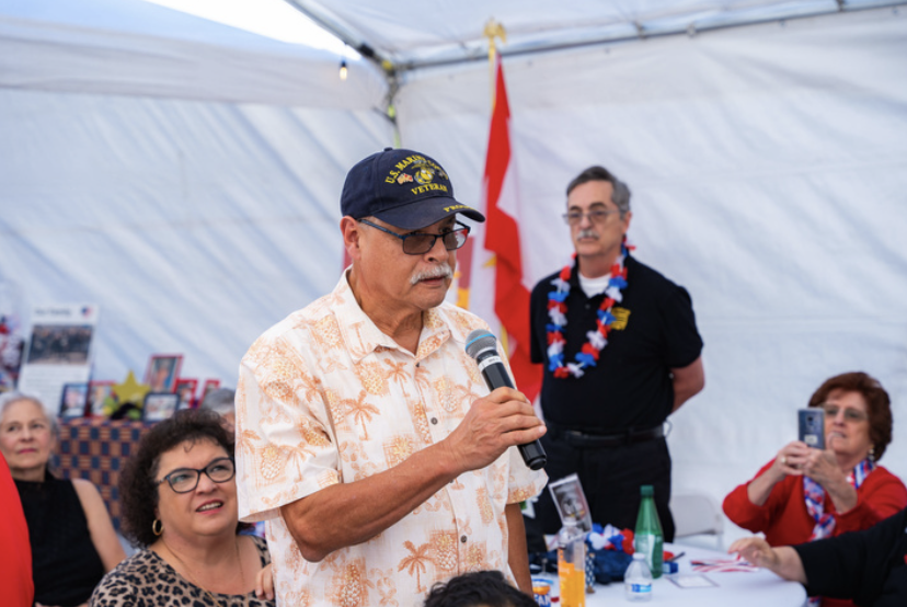 An elderly man with glasses, wearing a navy hat with military insignia and a tropical shirt, speaking into a microphone at a gathering inside a white tent decorated with American and military-themed items, with several seated and standing people list