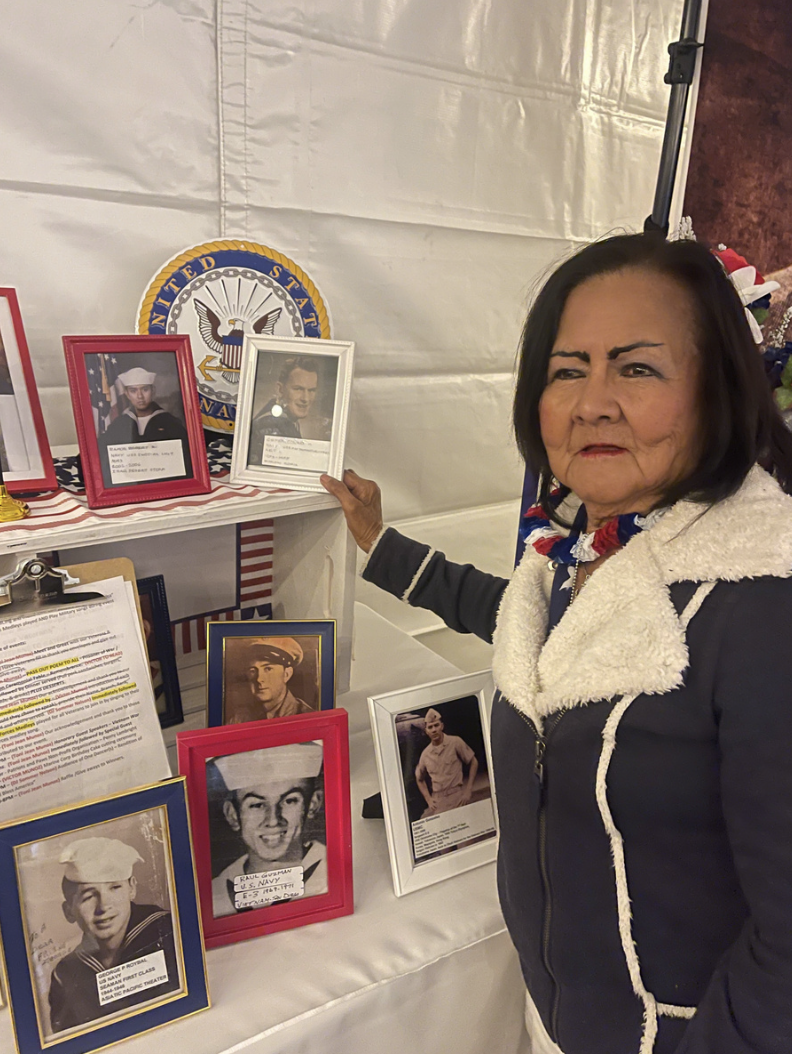 A woman standing next to a display of framed photographs of military personnel and a U.S. Navy emblem, in an indoor setting.