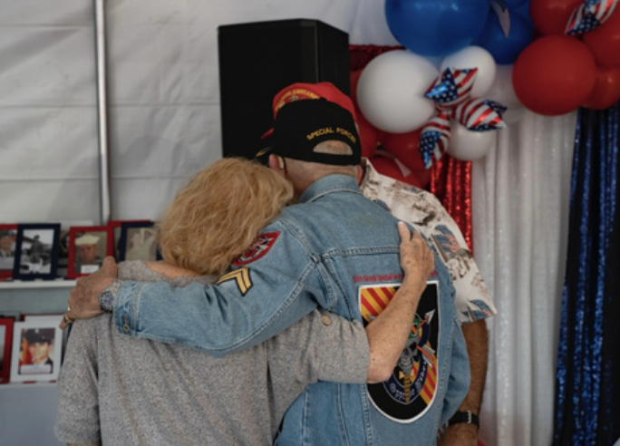 Three people hugging at a patriotic celebration, with red, white, and blue balloons and framed photos in the background.