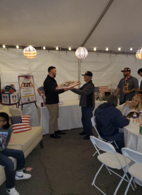 Man receiving a framed item from another man at a decorated tent with patriotic decor for a celebration, with people seated at tables and children present.