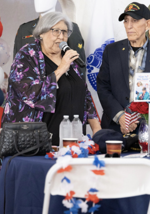 An elderly woman with gray hair and glasses speaking into a microphone at a patriotic event, surrounded by decorated tables and people wearing patriotic attire.