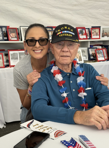 A smiling woman with sunglasses and bracelets stands behind an elderly man wearing a veteran hat, a patriotic lei, and a blue shirt, in front of a display of framed photographs.