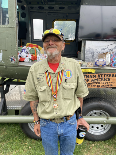 A happy man in a military veteran uniform with medals, standing in front of a military helicopter at an event, with various gifts and awards nearby.