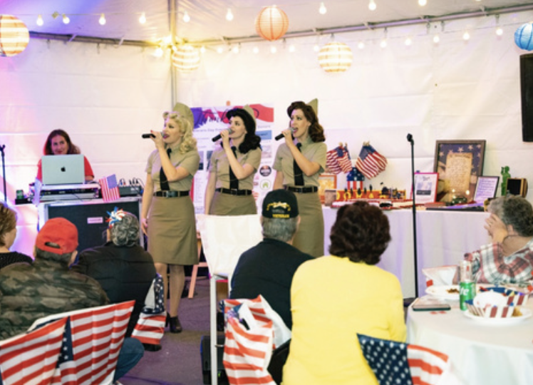 Three women singing into microphones at a patriotic-themed indoor event, with an audience seated at round tables decorated with American flags and patriotic colors.