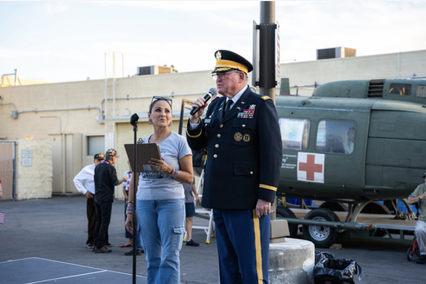 A man in a military dress uniform speaking into a microphone next to a woman holding a clipboard, with a green helicopter marked with a red cross in the background at an outdoor event.