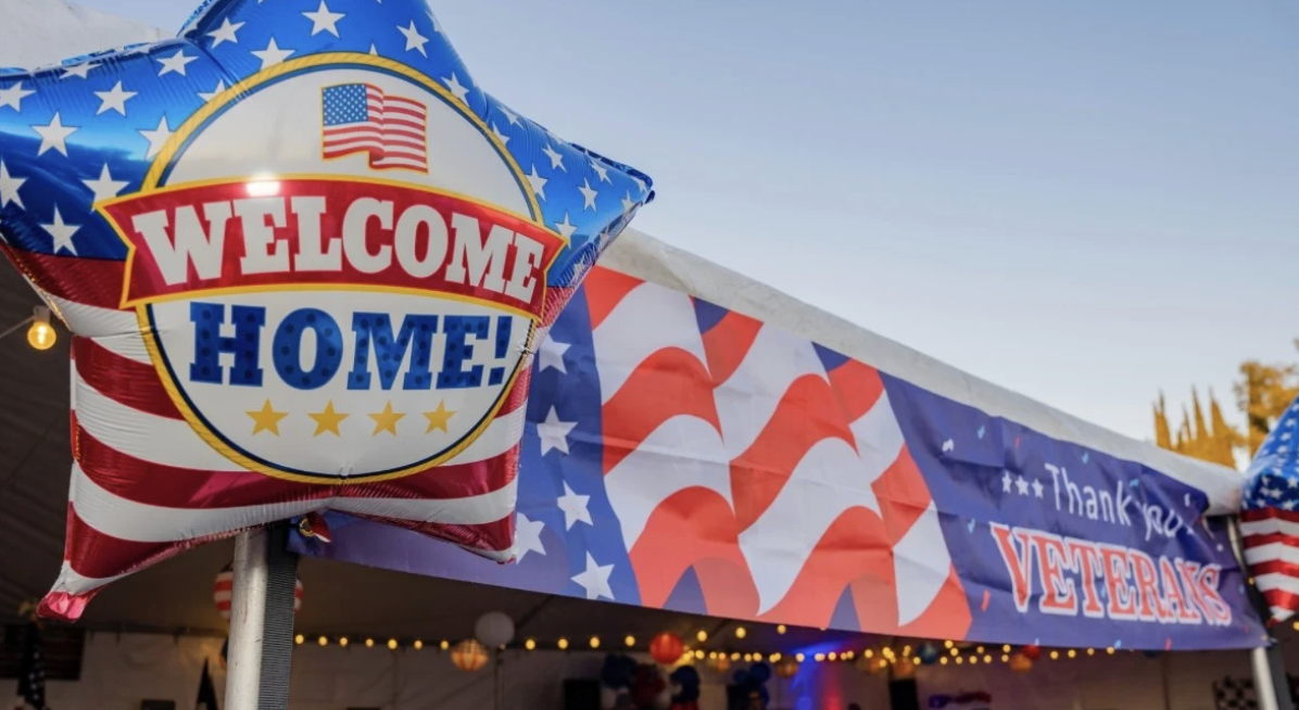 American-themed outdoor display with a balloon that says "Welcome Home!" and a banner thanking veterans, decorated with red, white, and blue colors and patterns.