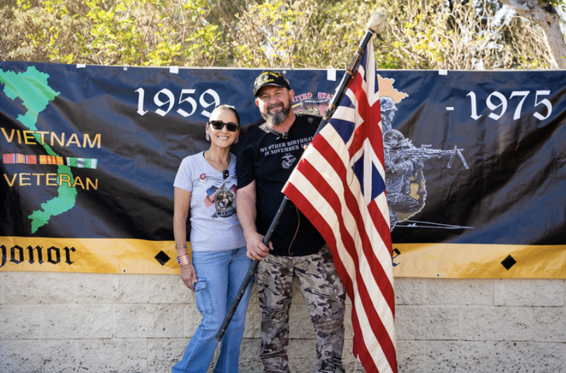 A man and woman standing outdoors in front of a Vietnam War veteran memorial banner. The man is holding an American flag, and both are smiling. The banner has the years 1959 and 1975 and features military imagery.
