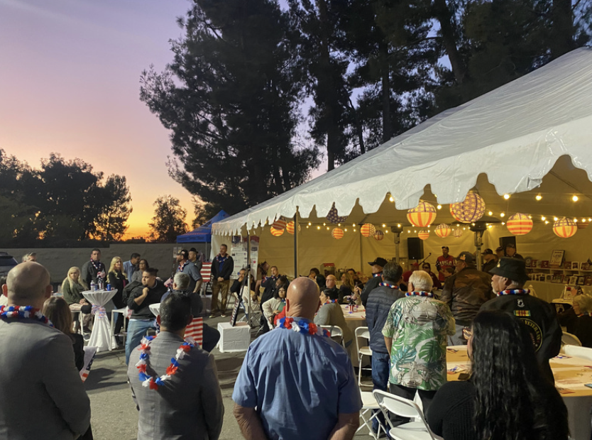 People gathered at an outdoor event during sunset, with a white tent decorated with orange and purple paper lanterns and string lights, featuring a live band or speakers in the background.