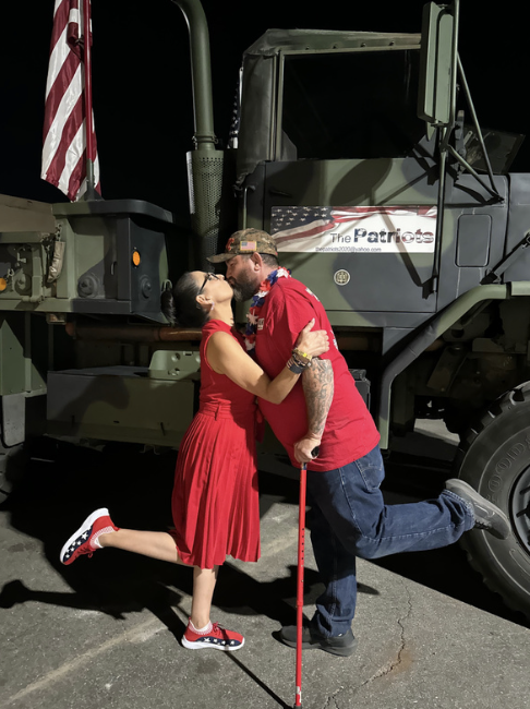 A couple in red clothing sharing a kiss in front of a military vehicle with American flags, one on the vehicle and one on a helmet, during a patriotic event.