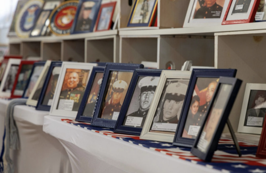Display of framed photographs of military personnel and veterans on a table and shelves, likely at a memorial or Veteran's event.