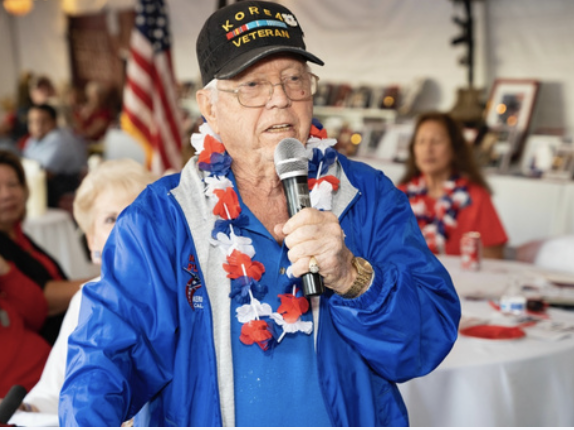 An elderly man speaking into a microphone at a gathering, wearing a black cap with Korean War Veteran and American flag-themed leis around his neck, with American flags and people in the background.