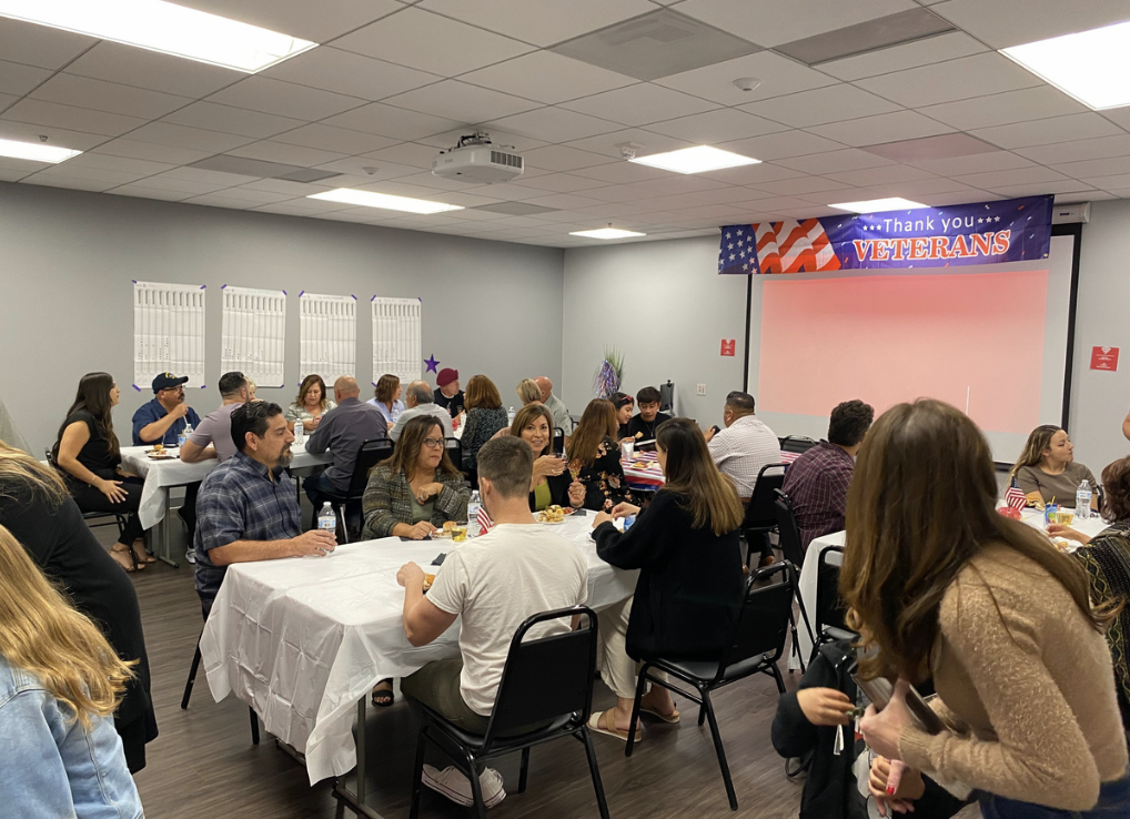 A group of people gathered in a room decorated to thank veterans, with tables, food, and a large screen, celebrating a veterans' appreciation event.