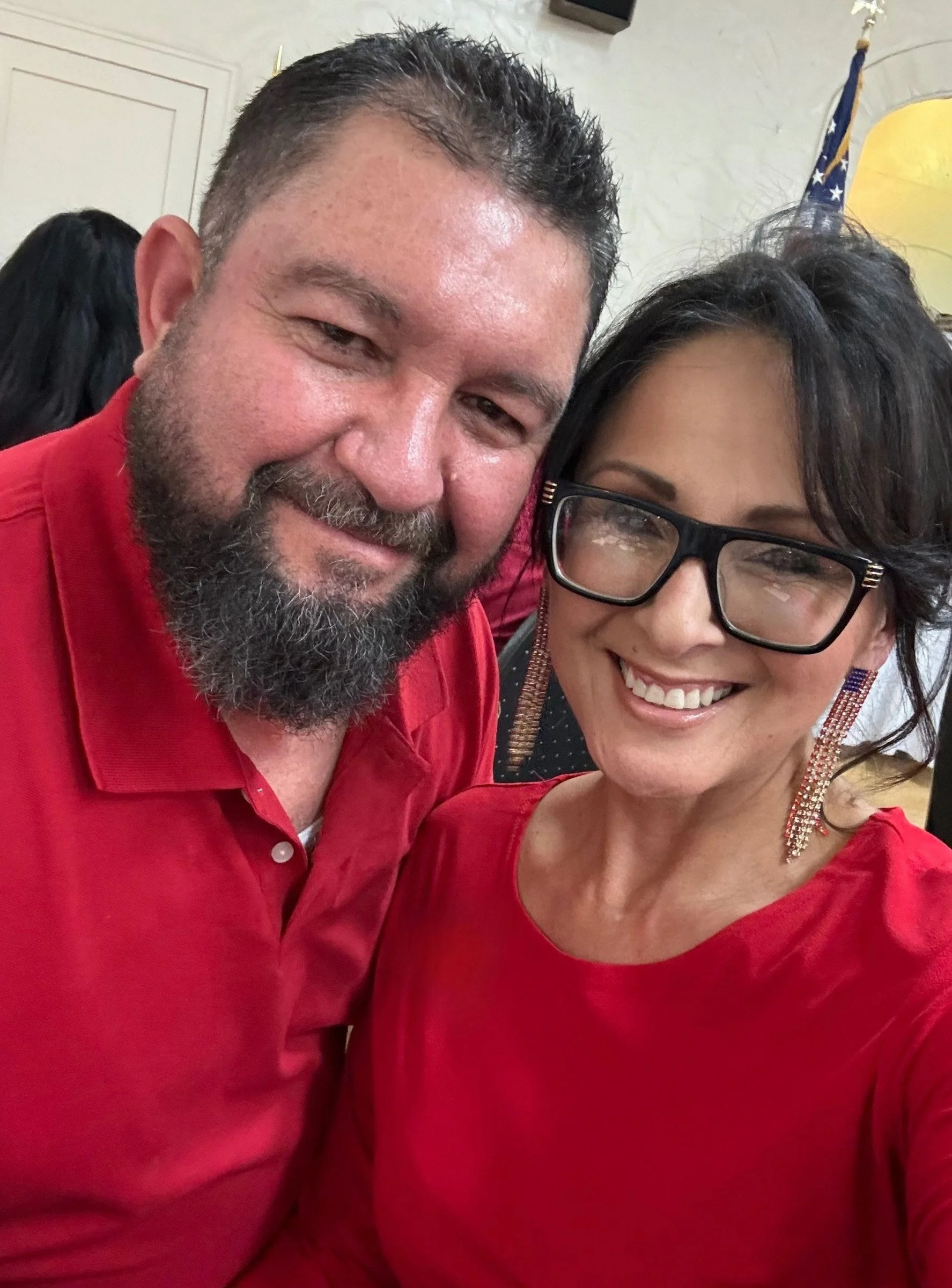 A close-up photo of a smiling man and woman, both wearing red tops, with the man having a beard and short hair, and the woman wearing glasses and earrings, in an indoor setting with an American flag and a lamp in the background.