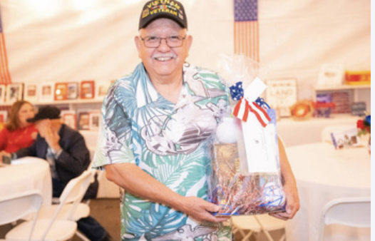 Older man smiling and holding a gift basket decorated with an American flag ribbon at a patriotic event with a U.S. flag in the background.