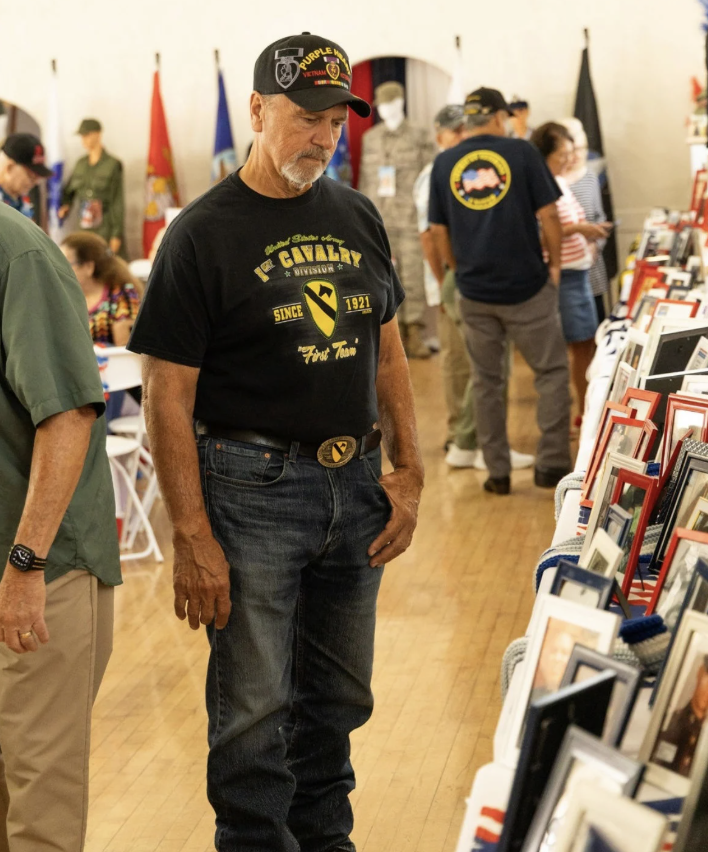 An older man wearing a black 'Vietnam Veteran' cap and a black T-shirt with military-related text and insignia, standing and looking at picture frames on display at an indoor event.
