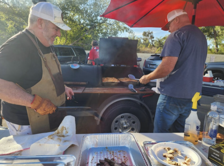 Two men cooking barbecue near a parked truck with a red umbrella providing shade, with various food trays and condiments on the table in front of them.