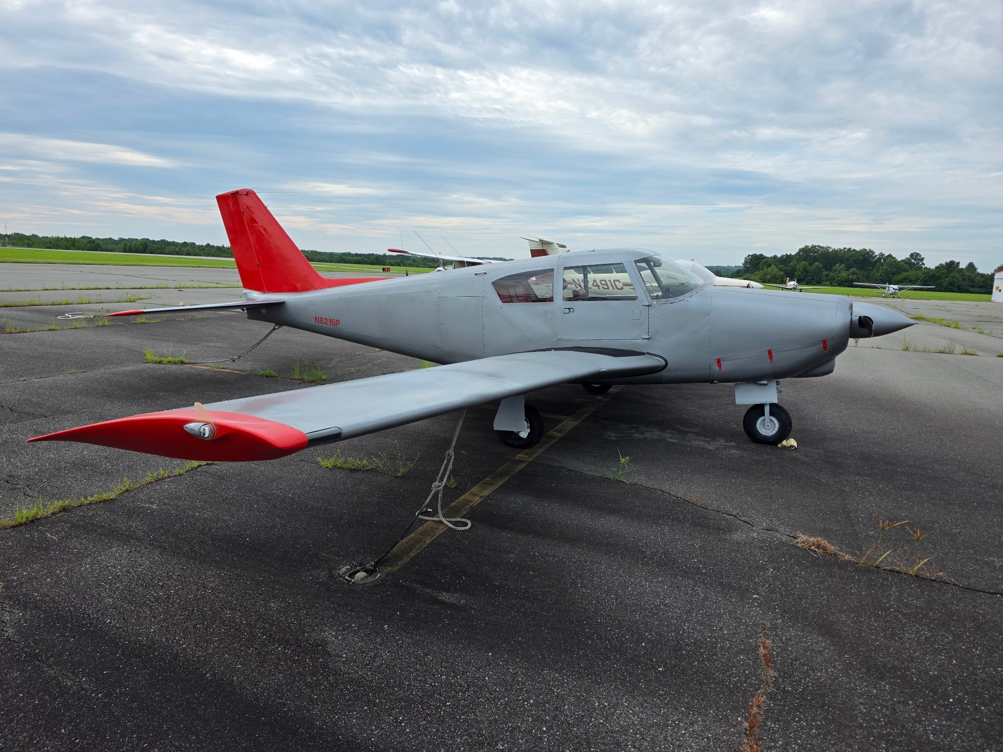 A small gray aircraft with a red tail and red accents on the wings and nose parked on an airport tarmac. The background shows other small airplanes parked on the grassy field under a cloudy sky.