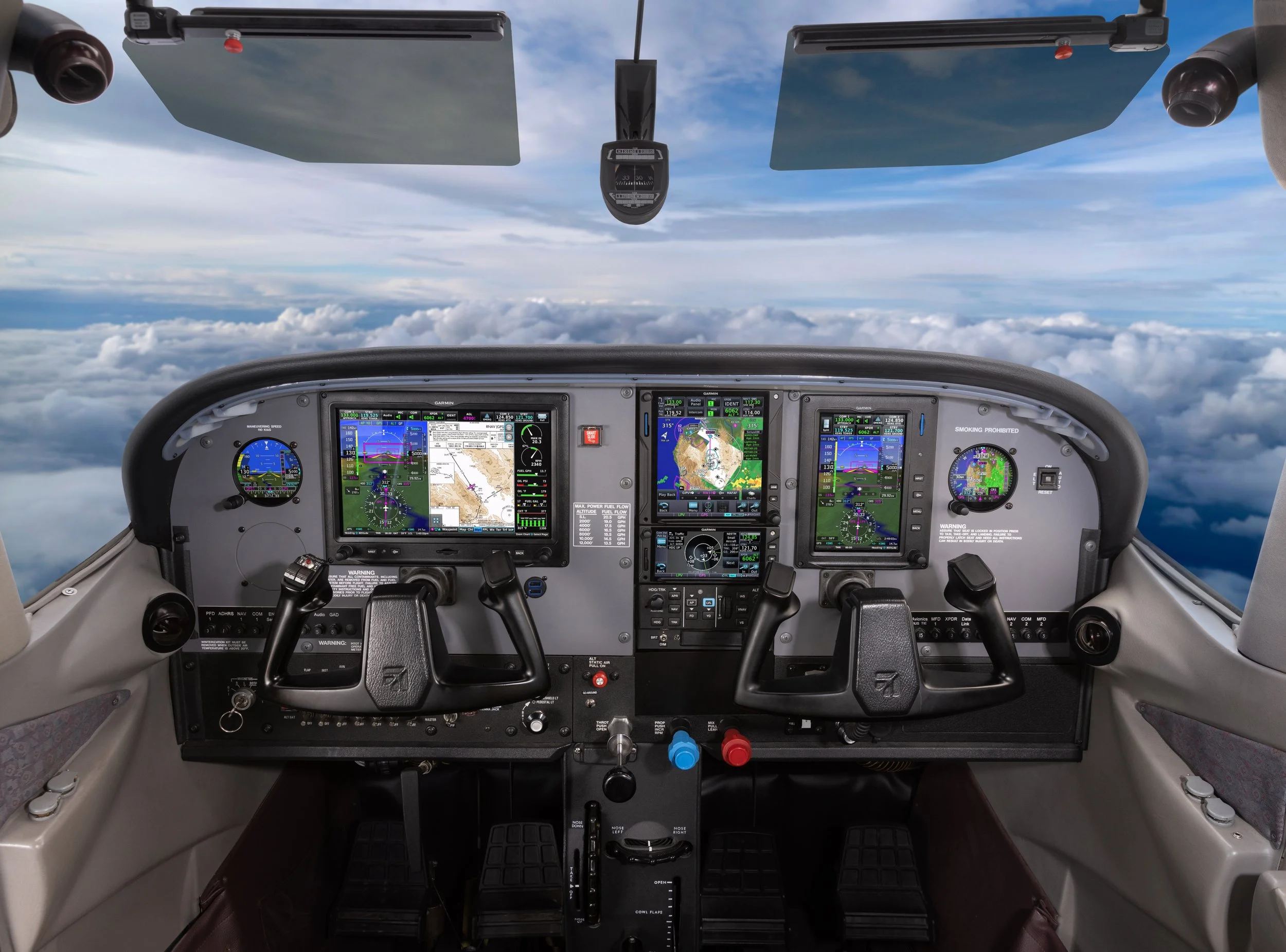 Inside view of an airplane cockpit showing various digital instruments and gauges, with clouds and sky visible through the windshield.