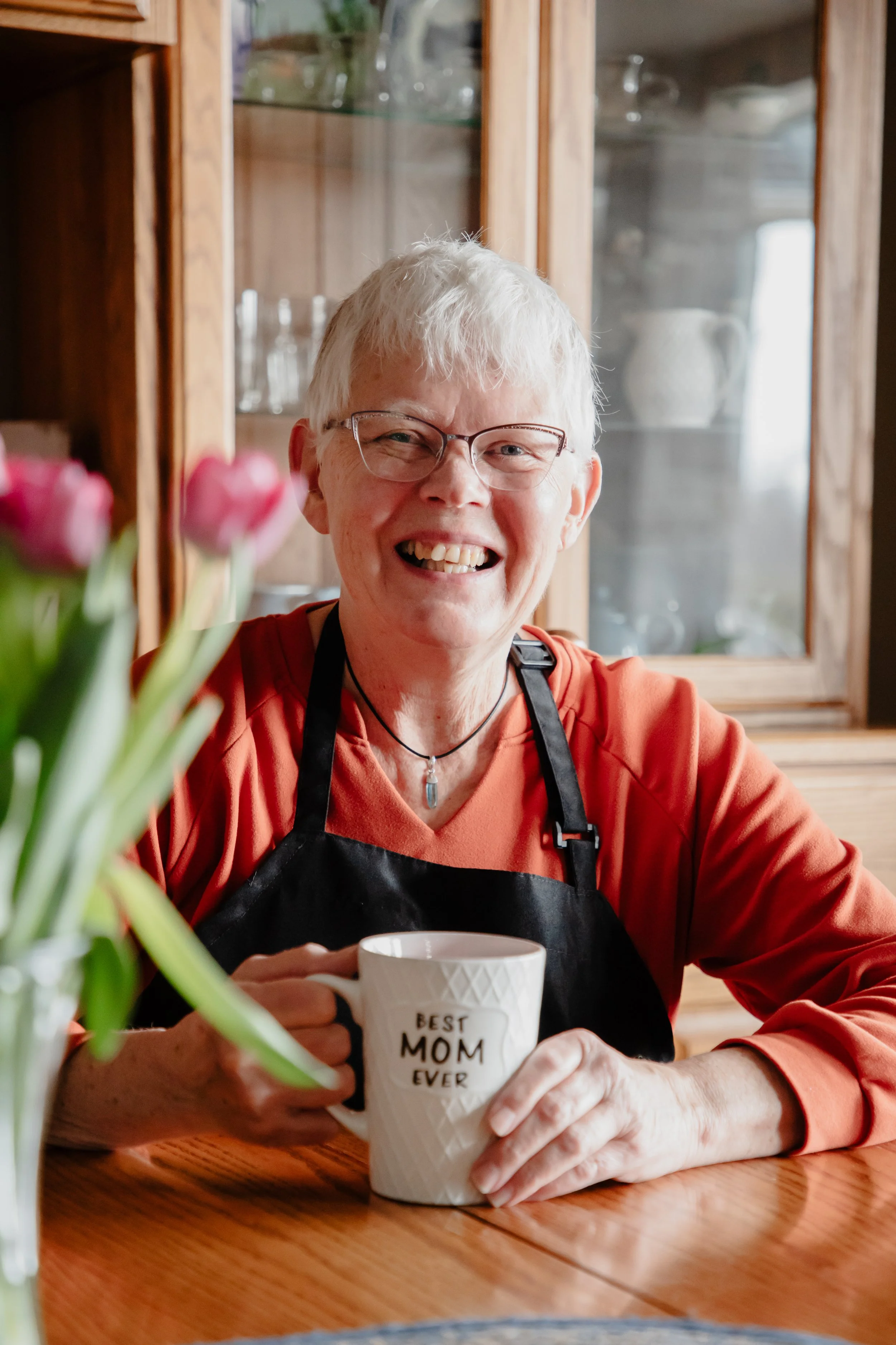 A woman with short gray hair, glasses, and a red shirt, wearing a black apron, sitting at a wooden table, holding a white mug that says "Best Mom Ever," smiling at the camera, with a glass-fronted cabinet in the background and pink tulips in a vase on the table.