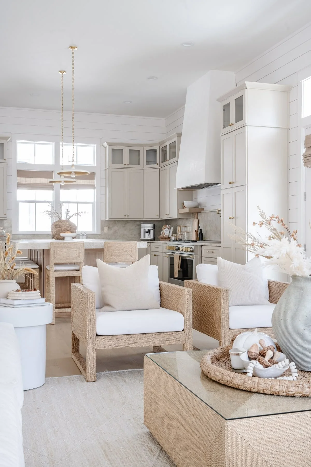 A bright, airy kitchen and living room area with neutral, light-colored furnishings and decor. Includes white cabinetry, a large window with natural light, rattan chairs, a white sofa with pillows, and a beige textured coffee table with decorative items. The space has a coastal and minimalist aesthetic.