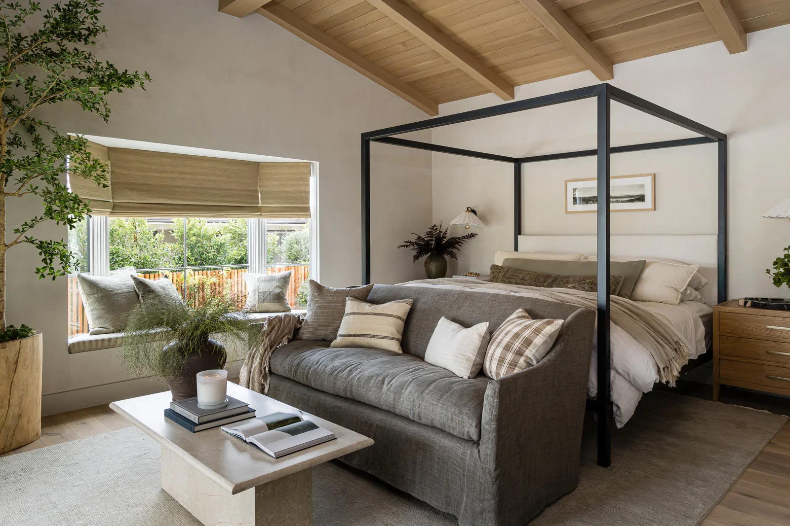 A bedroom with a four-poster bed, a gray sofa with pillows, a large window with blinds, and a coffee table with books and decorative items, in a room with wooden ceiling beams and a potted plant.